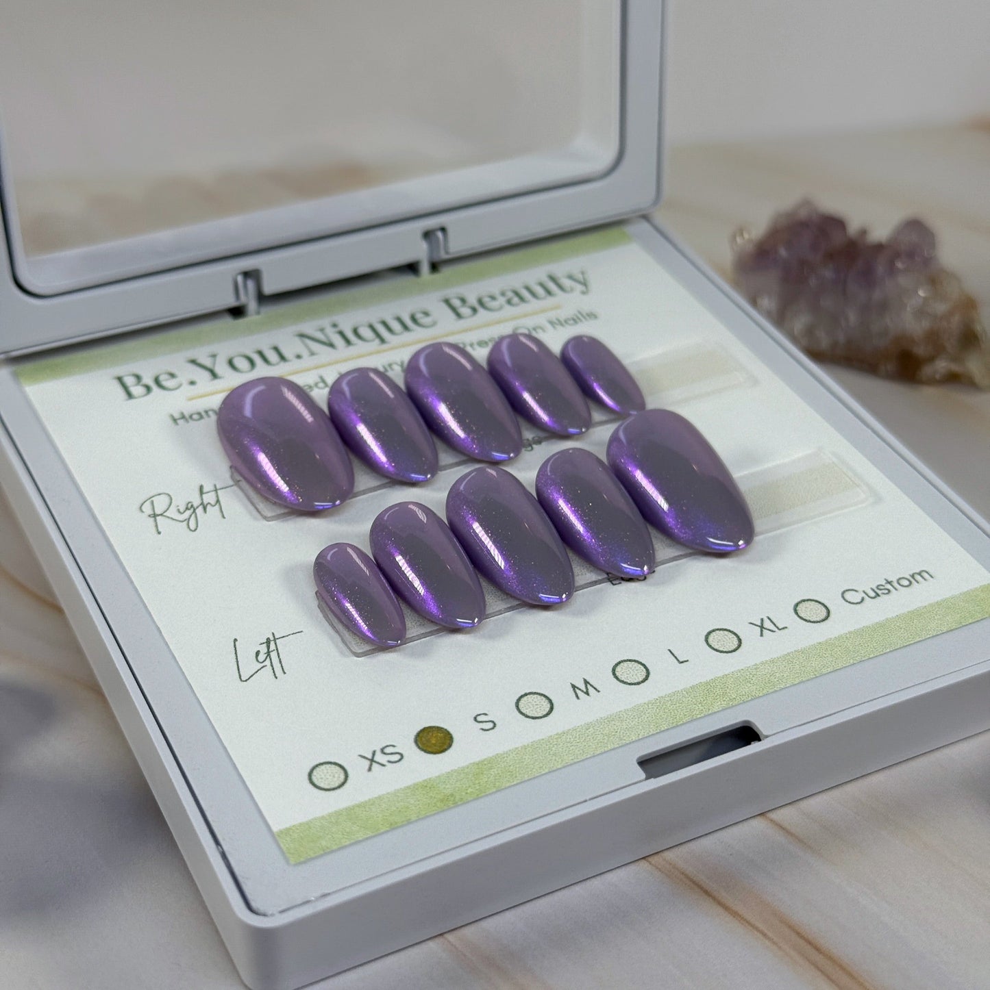 A set of purple chrome press on nails set in a branded case on a marble surface