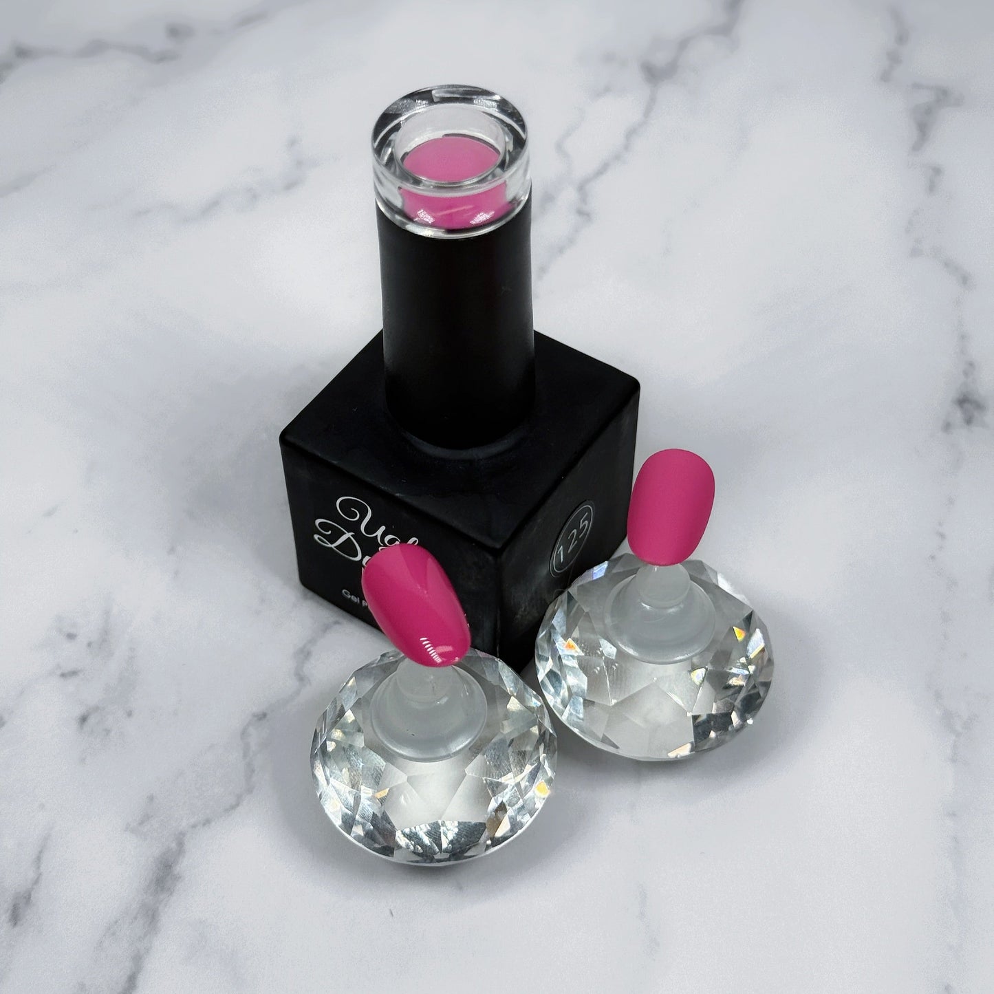 Nail polish bottle with pink top and two sample nails showing the colour with a matte and glossy topcoat on a marble surface.
