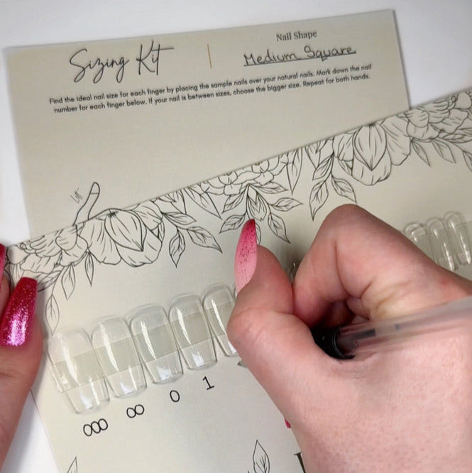 A card with clear nail tips in medium square with a hand wearing pink sparkly ombre press on nails is writing the corresponding nail numbers under them for a customer to use to size their natural nails for correct fitting press ons.