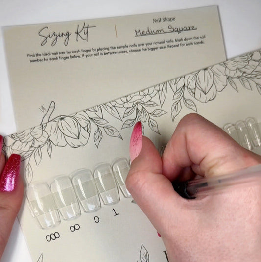 A card with clear nail tips in medium square with a hand wearing pink sparkly ombre press on nails is writing the corresponding nail numbers under them for a customer to use to size their natural nails for correct fitting press ons.