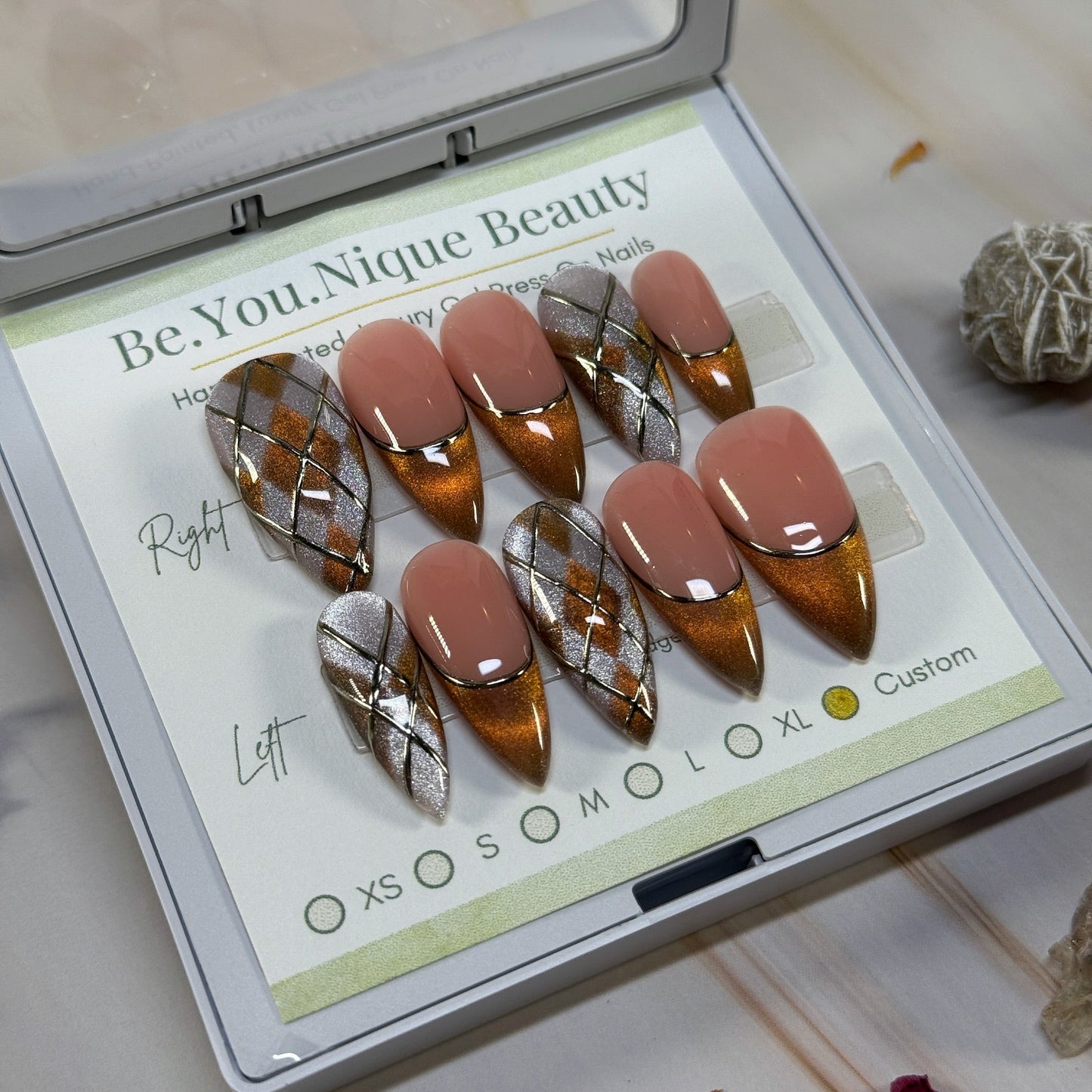 Set of false nails with an argylle and french tip design in gold and pink cat eye with gold chrome accents in a display case with decorative stones and crystals on a marble surface.