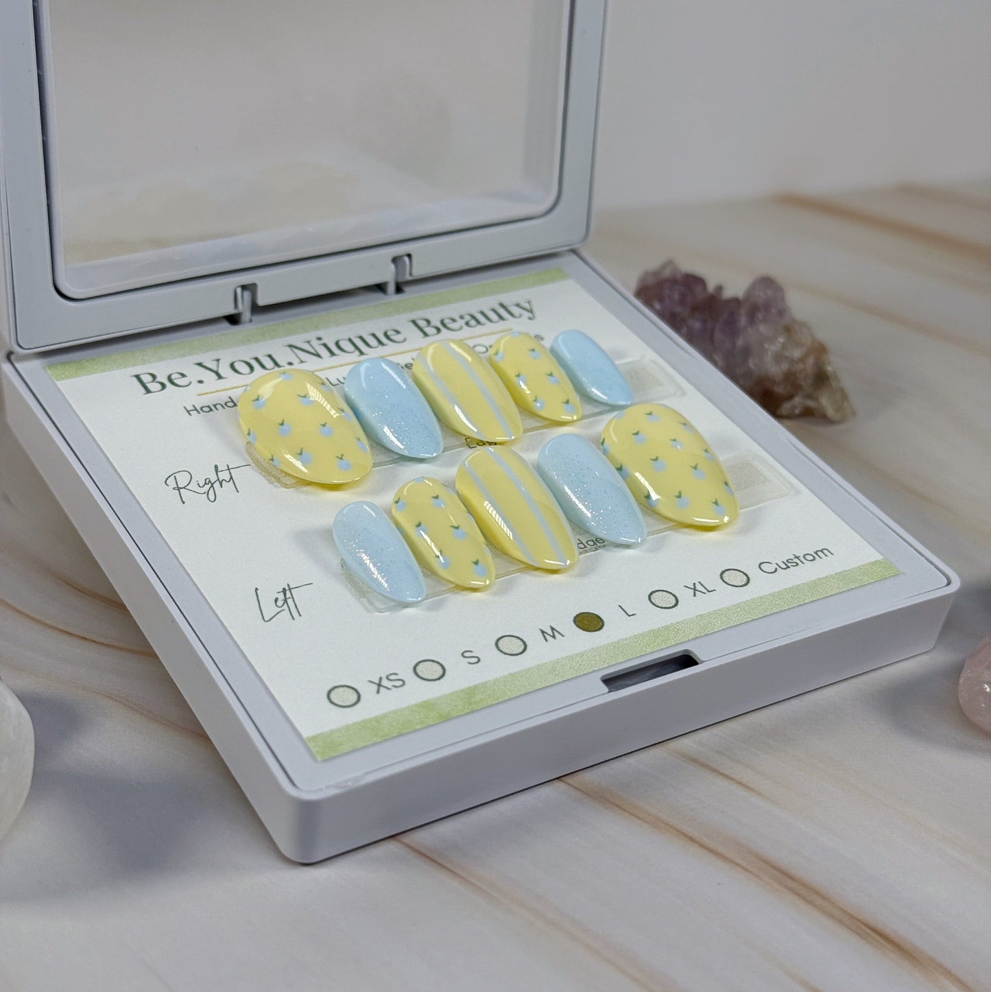 Set of yellow and blue nail art designs in a 'Be.You.Nique Beauty' box on a marble surface.