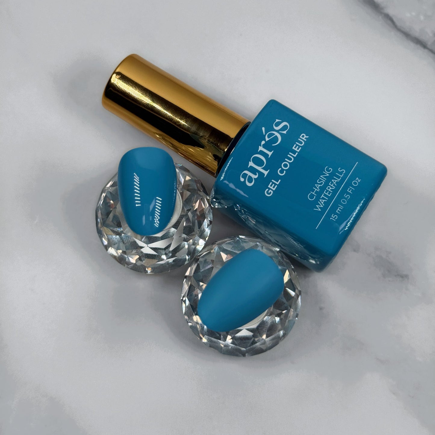 Blue nail polish bottle with gold cap on a marble surface with two sample nails showcasing the colour with a matte versus glossy topcoat.