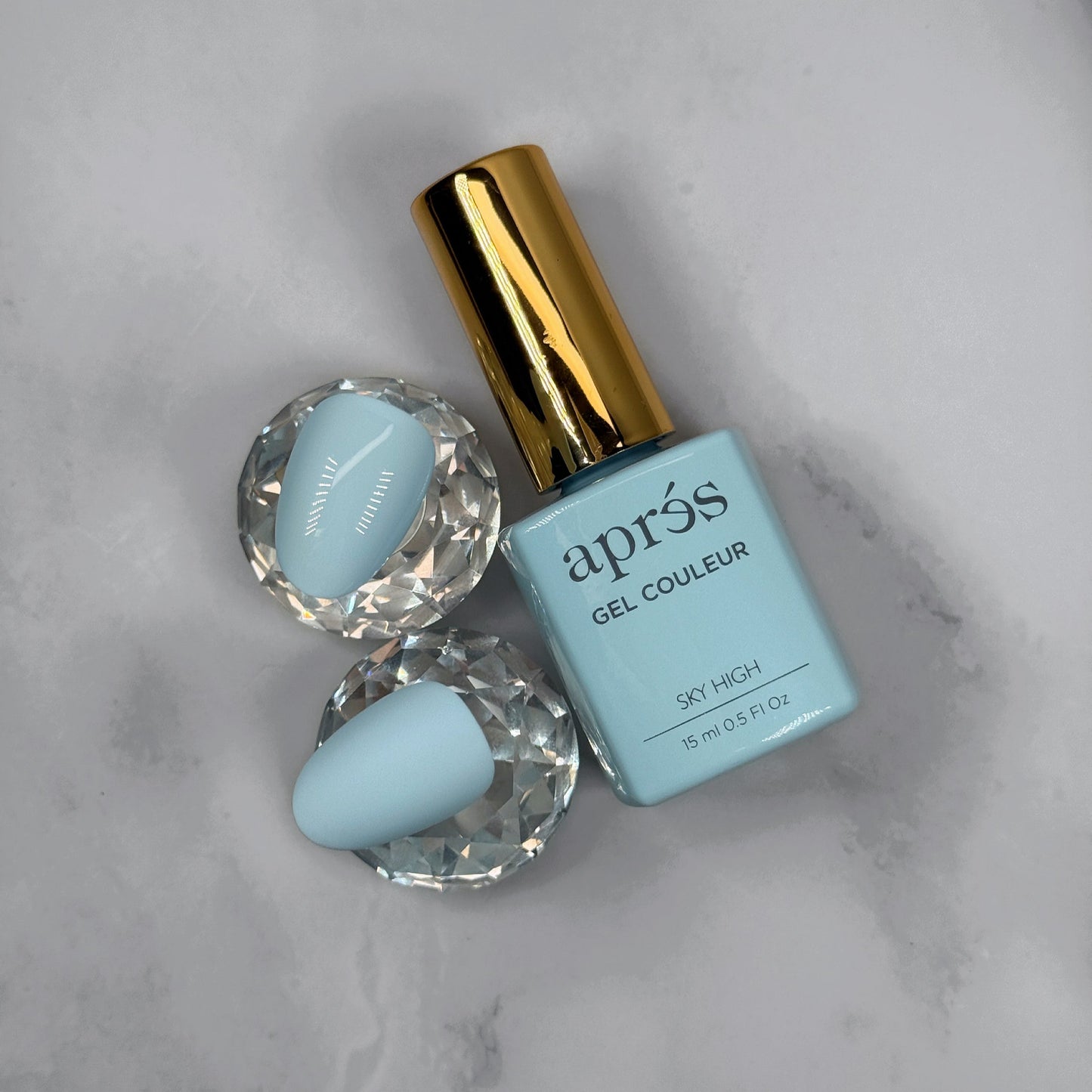 Blue nail polish bottle with gold cap on a marble surface and two sample nails showing glossy versus matte topcoat