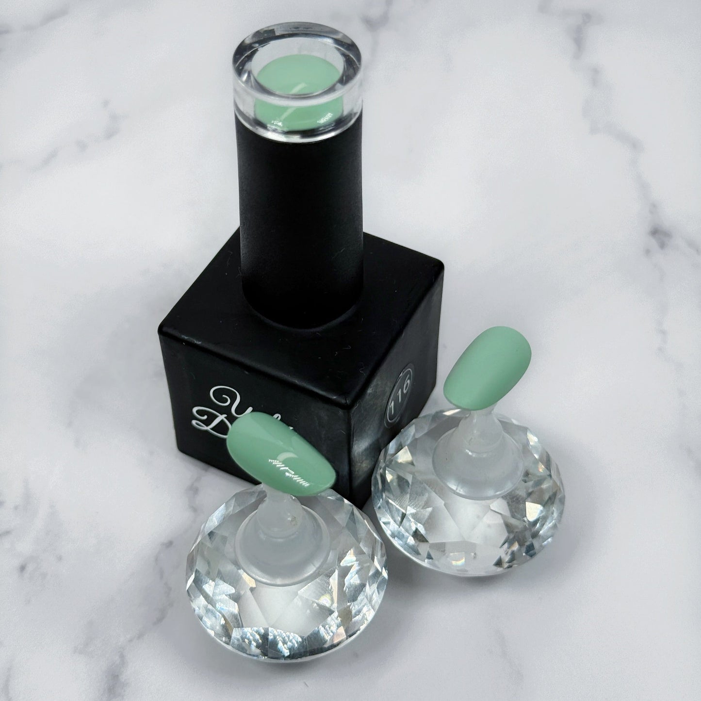 A nail polish bottle with two sample nail tips showing a light mint/sage tone. Each nail shows the difference between matte and glossy topcoats.