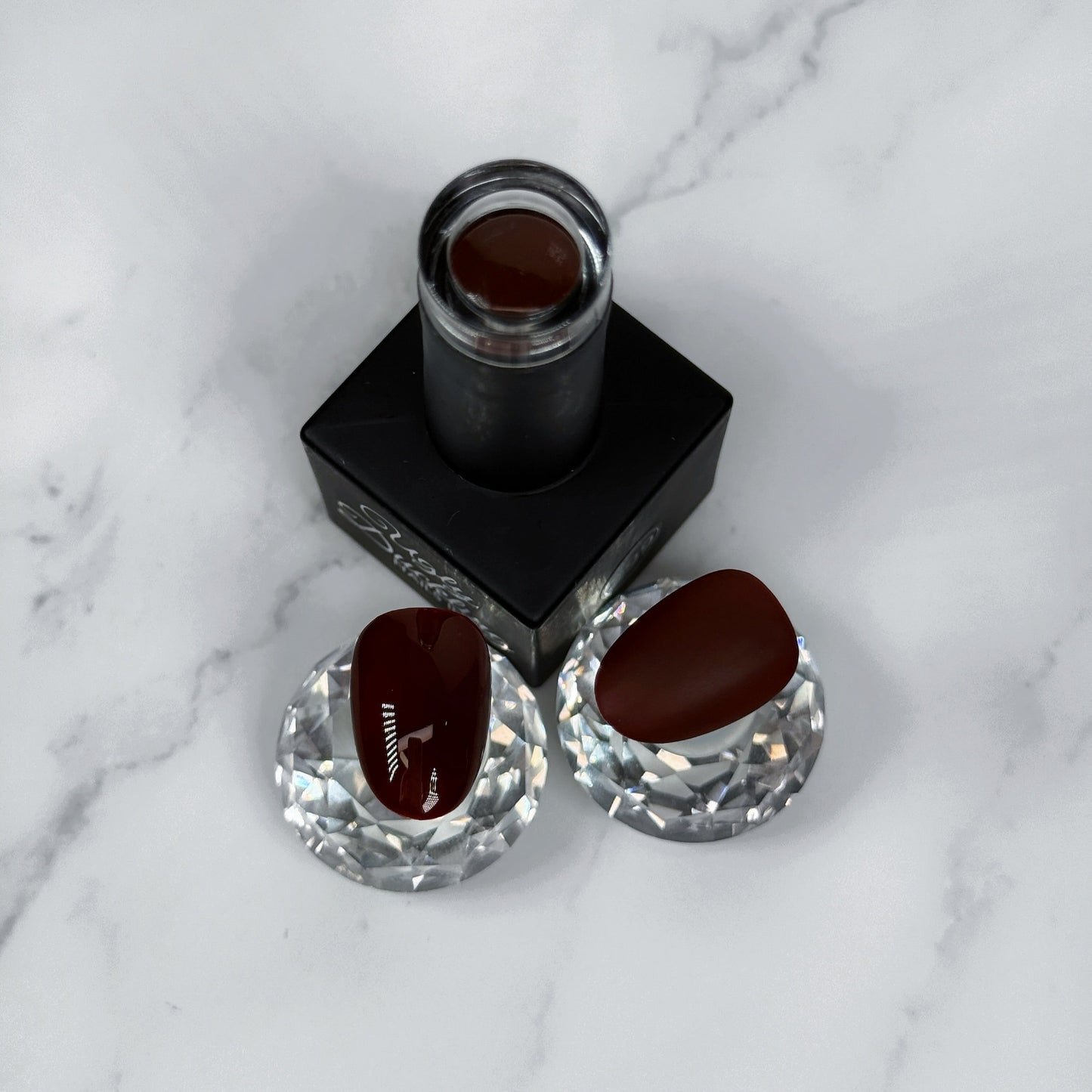 Nail polish bottle with two sample nails showing the brown colour with a glossy and matte topcoat on a marble surface