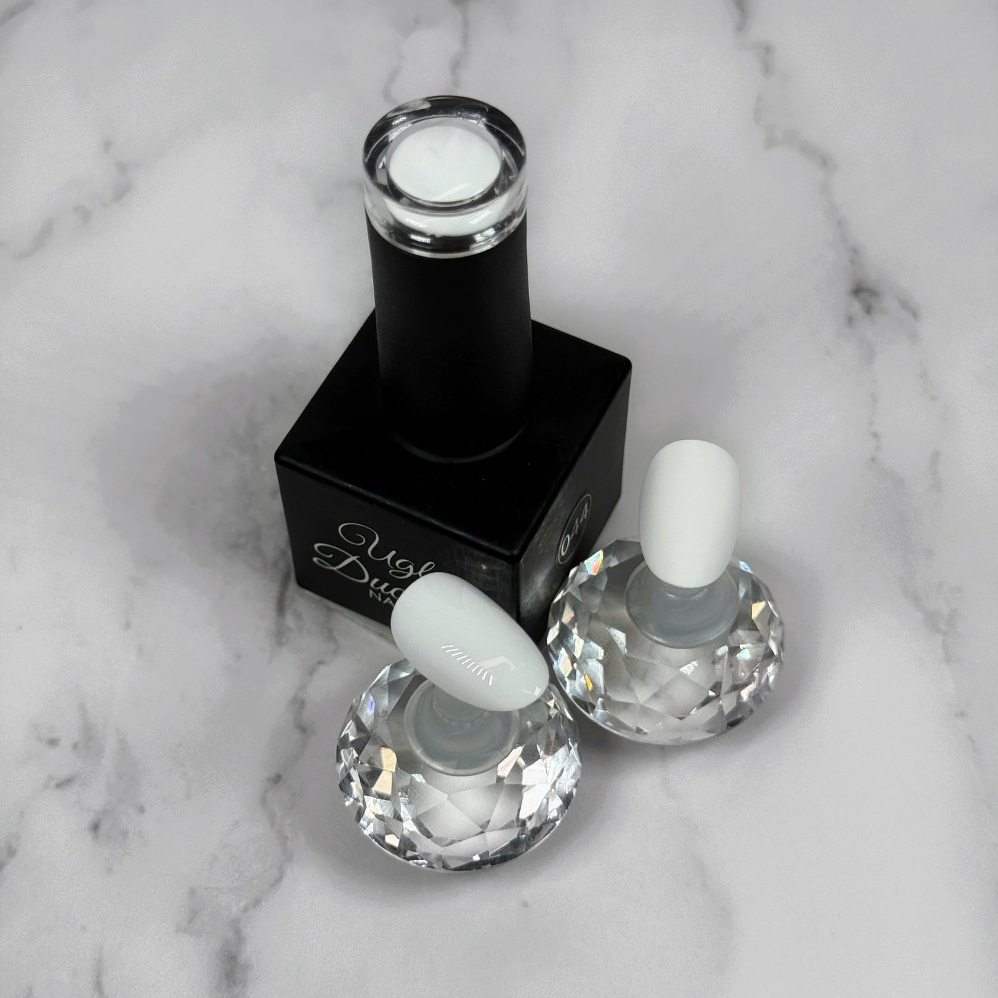 Black nail polish bottle with two nail samples showing glossy and matte topcoat on a marble surface