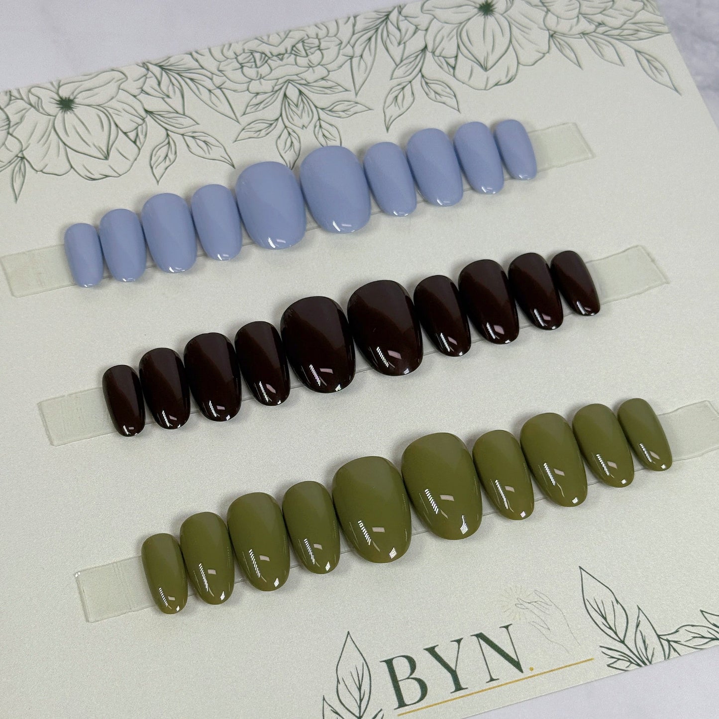 3 sets of press on nails in blue, brown, and green on a branded card with floral designs.