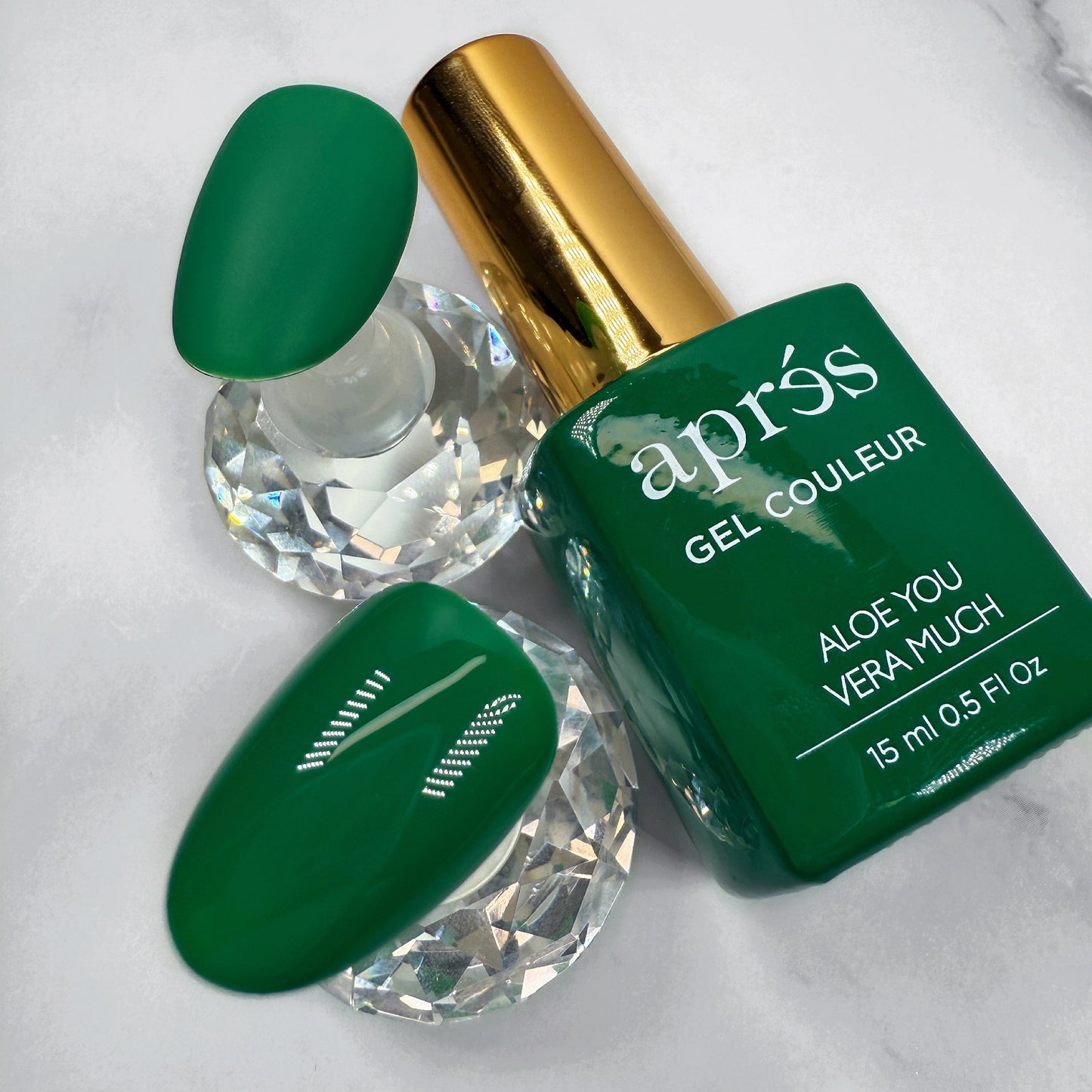 Green nail polish bottle with gold cap on a white marble surface with two samples nails showing matte and glossy topcoat