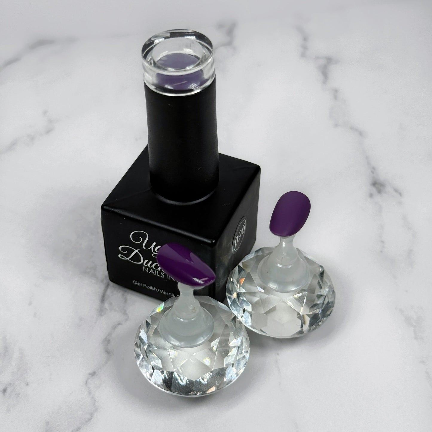 Black nail polish bottle with purple cap  and two sample nails showcasing the colour with either matte or glossy topcoat on a marble surface