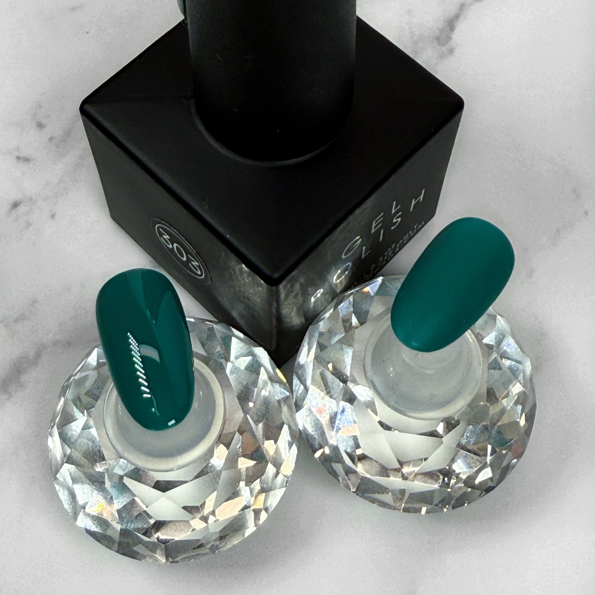 Teal nail polish bottle with two matching nail samples showing a gloss and matte topcoat option on a marble background