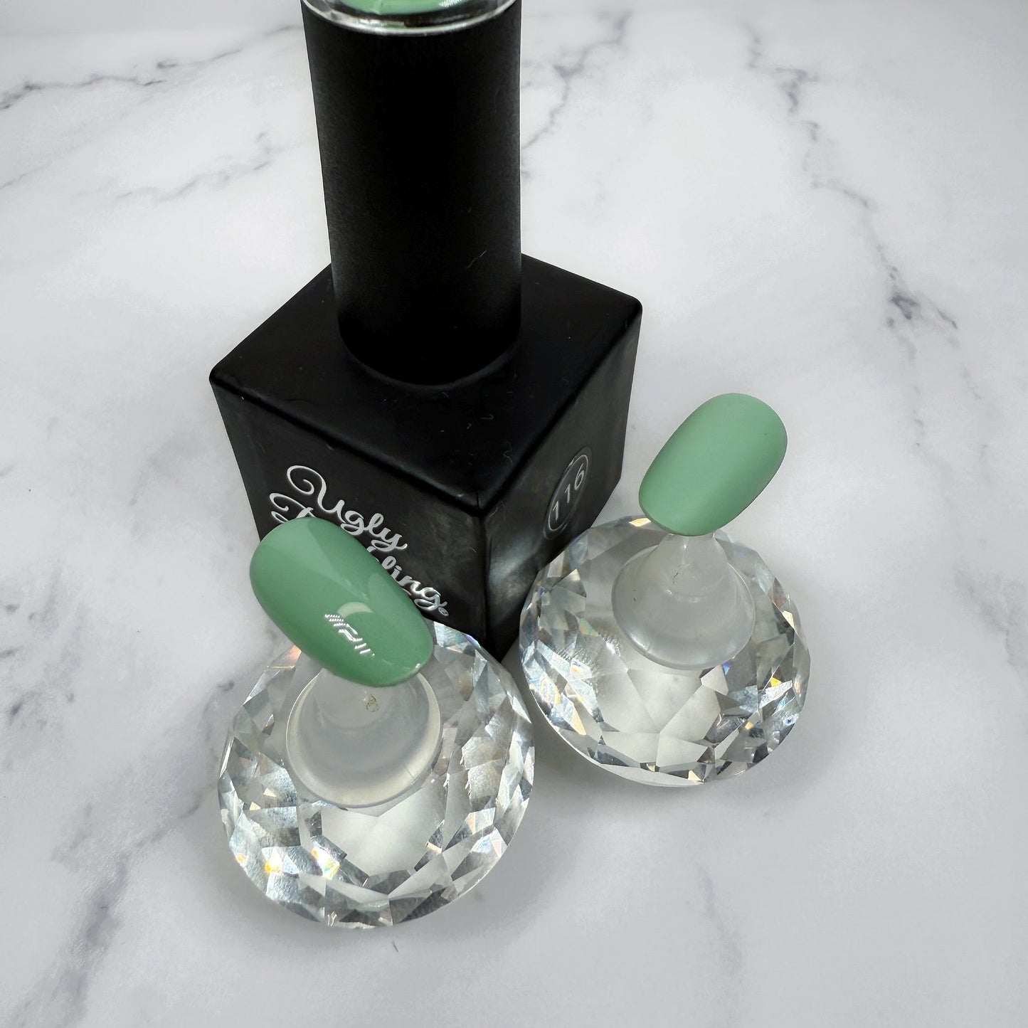 A nail polish bottle with two nail tips showing the light mint/sage colour with the difference between matte or glossy topcoat.