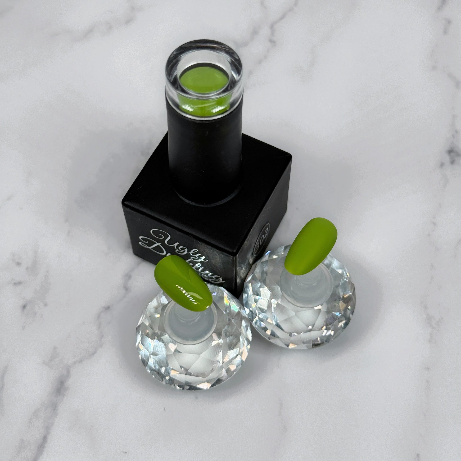 A nail polish bottle with two sample tips in front of it showcasing the polish colour with a matte and glossy topcoat.
