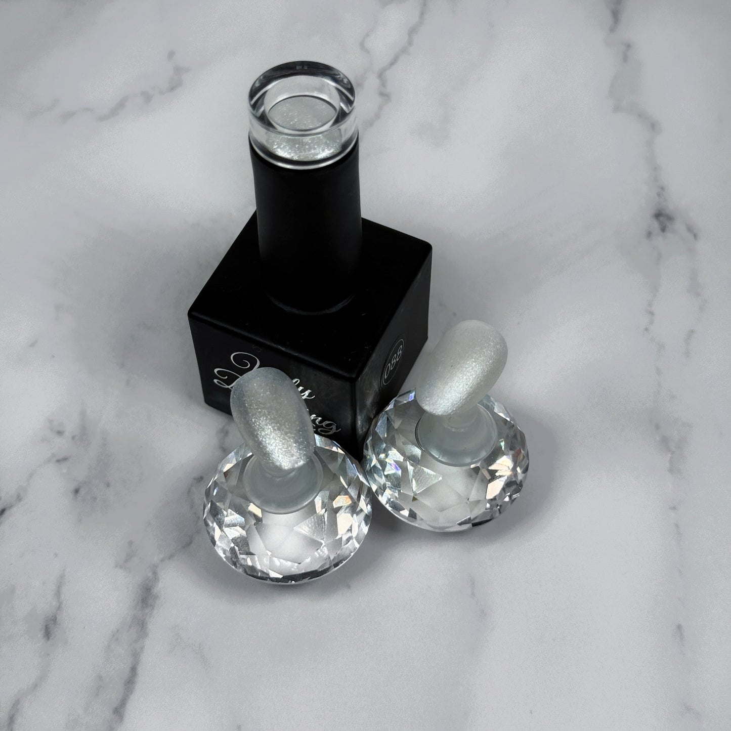black nail polish bottle with two sample nails showing a shimmery white polish with a glossy and matte topcoat on a marble background