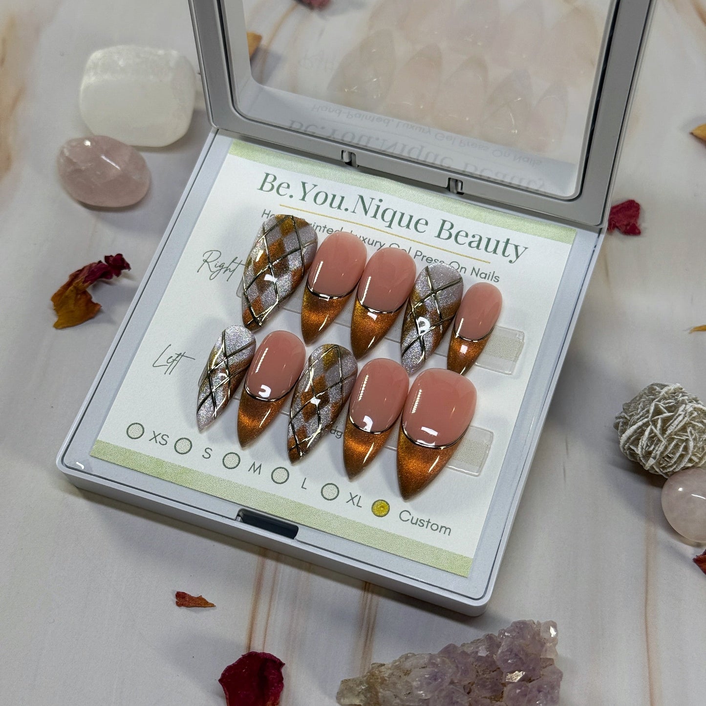 Set of false nails  with an argylle and french tip design in gold and pink cat eye with gold chrome accents in a display case with decorative stones and crystals on a marble surface.