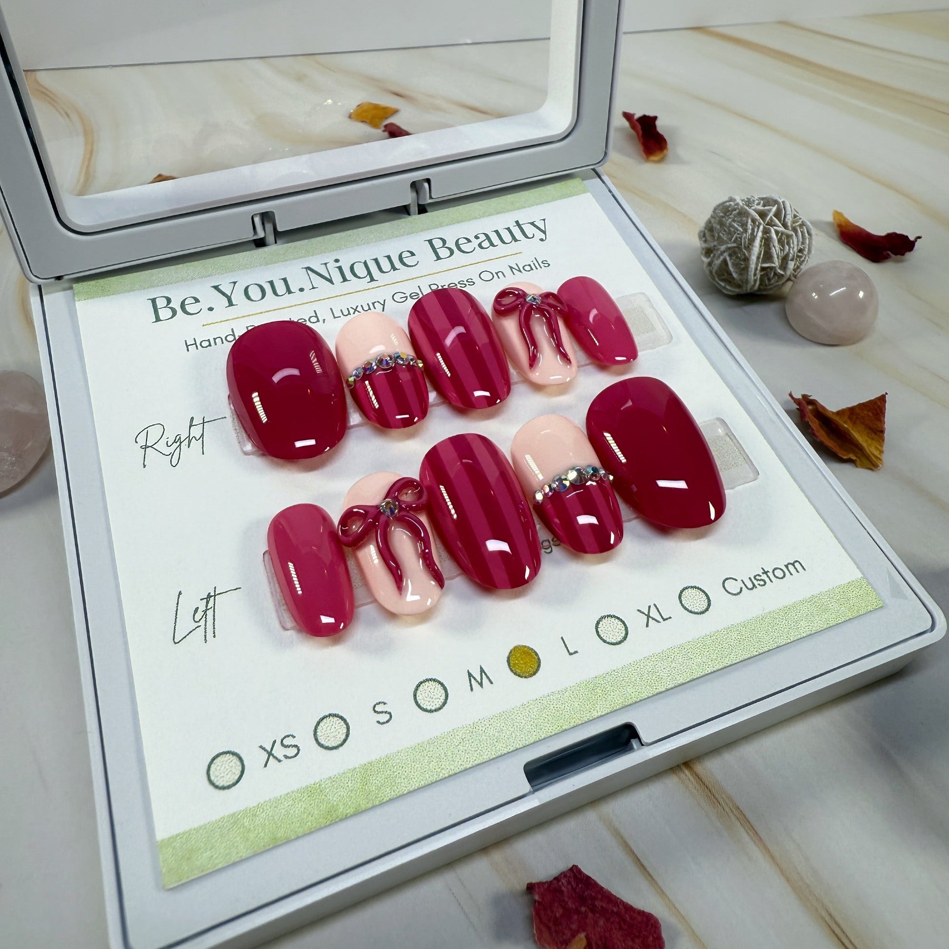 Set of Maroon and pink with stripes, gems, and a 3D bow on false nails in a display case with 'Be.You.Nique Beauty' branding.