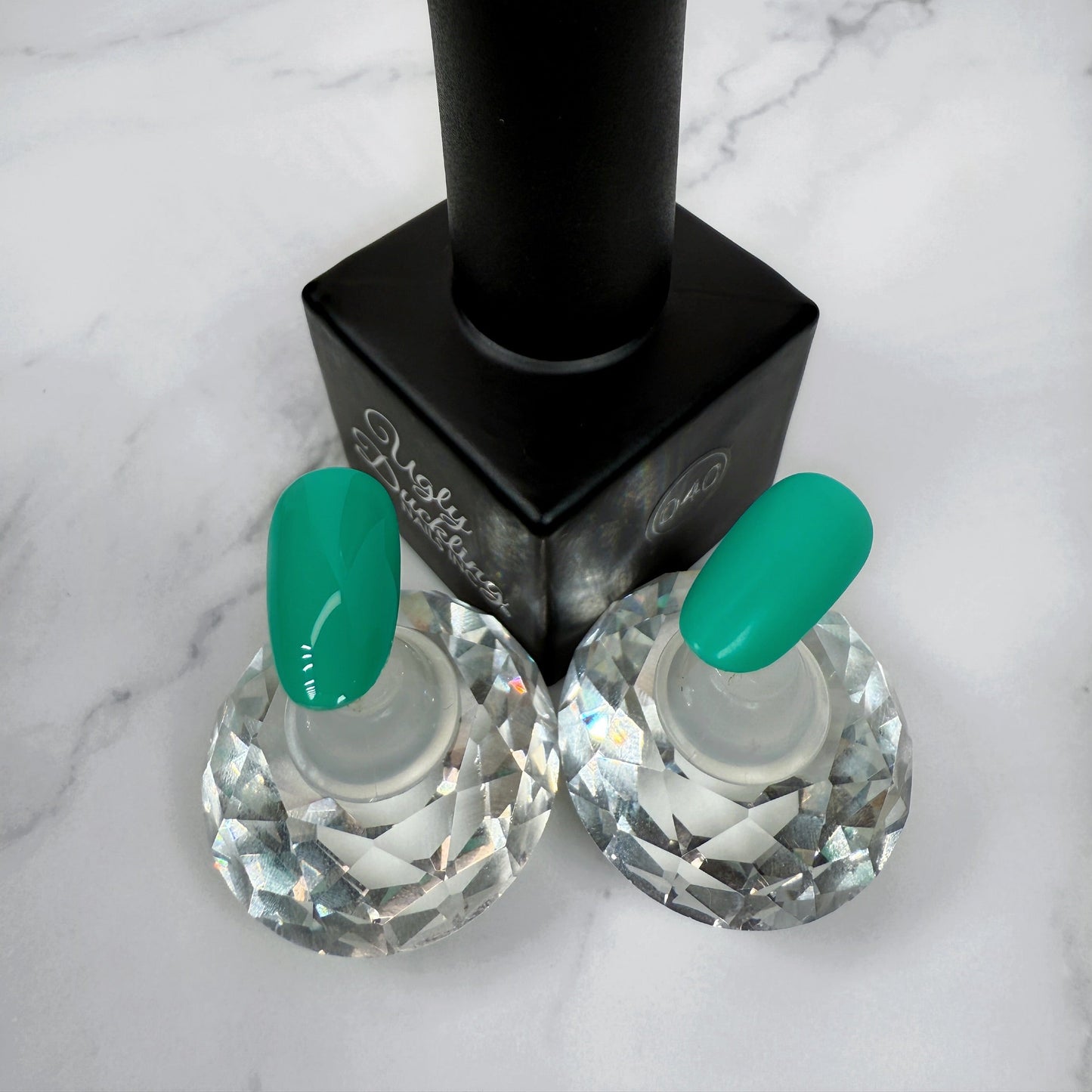 a nail polish bottle with two nail tips showcasing a light teal colour, the two nails show the difference between matte and glossy topcoats.