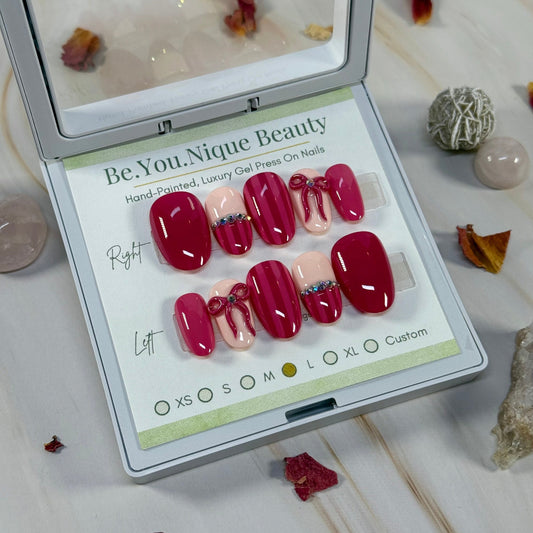 Set of Maroon and pink with stripes, gems, and a 3D bow on false nails in a display case with 'Be.You.Nique Beauty' branding.