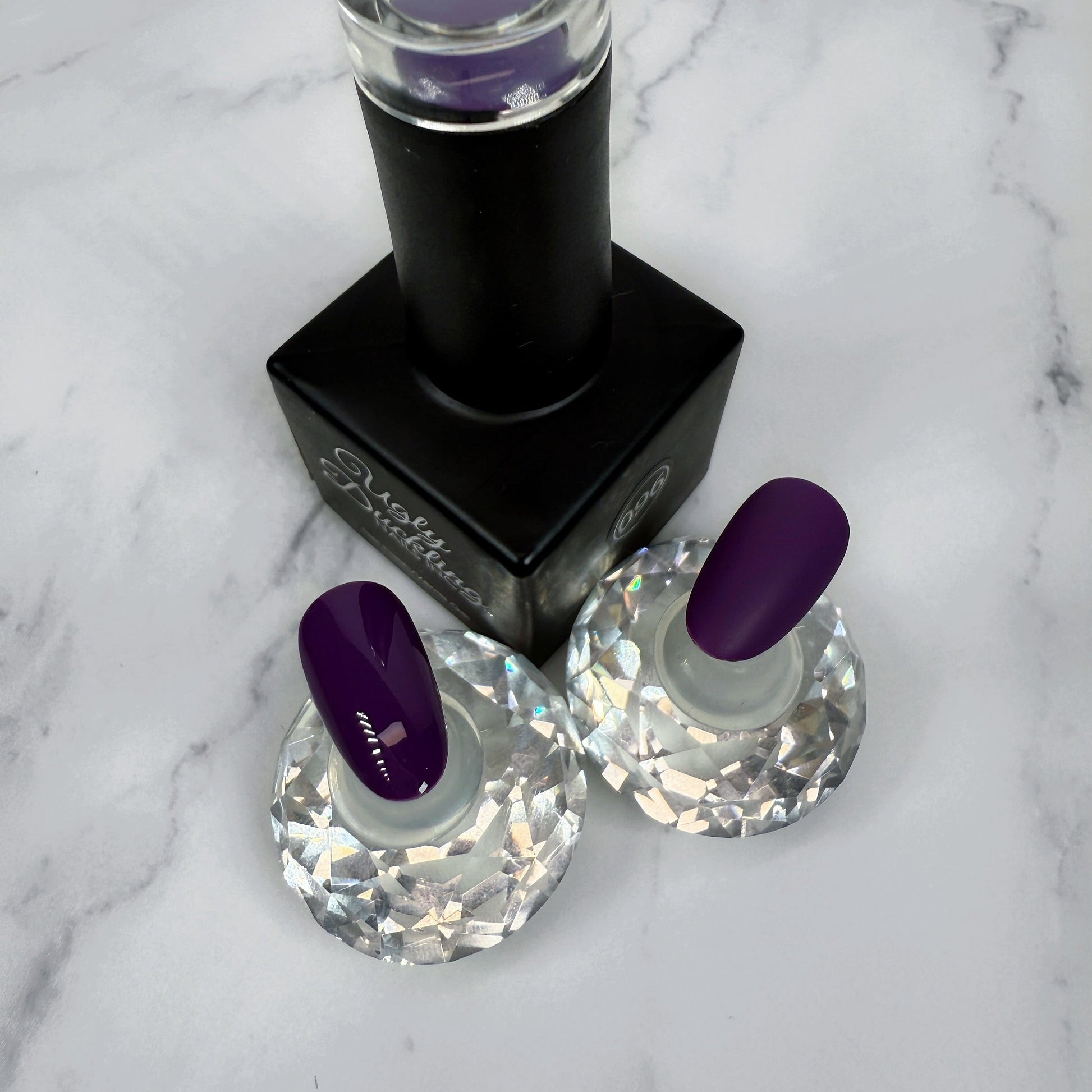 nail polish bottle with two sample nails showcasing a deep royal purple polish with a glossy and matte topcoat option.