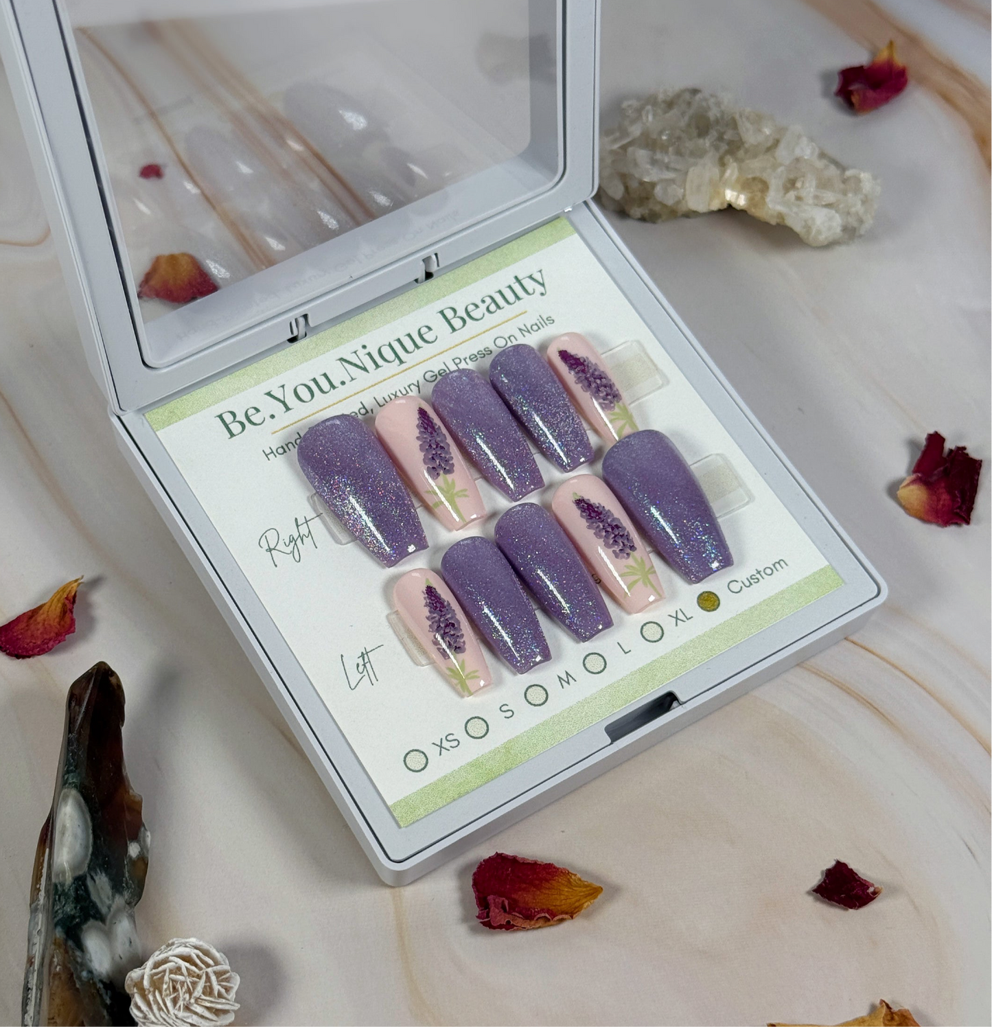 Set of purple and pink press-on nails in a 'Be.You.Nique Beauty' box on a decorative surface with crystals and flowers.