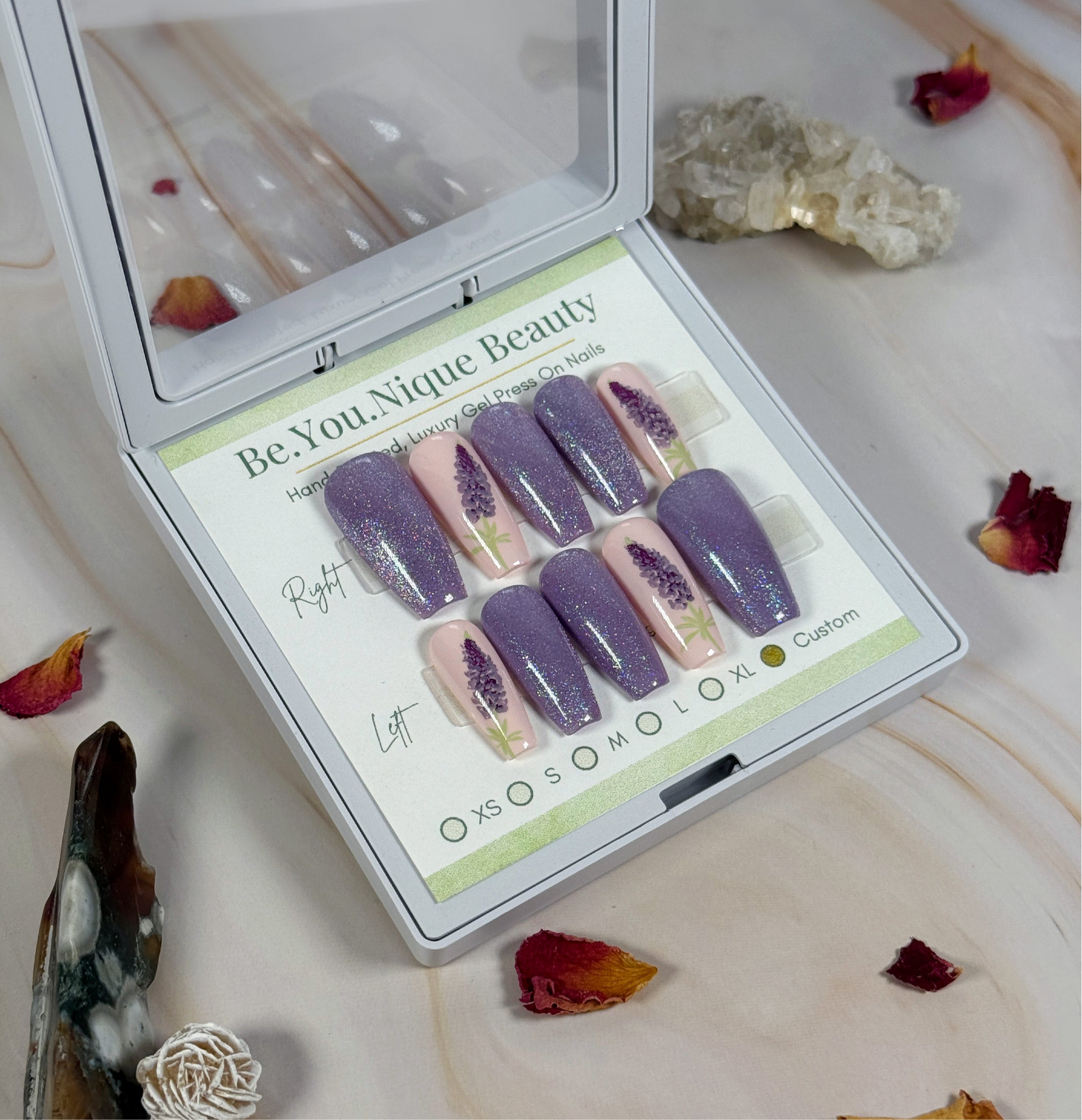 Set of purple and pink press-on nails in a 'Be.You.Nique Beauty' box on a decorative surface with crystals and flowers.