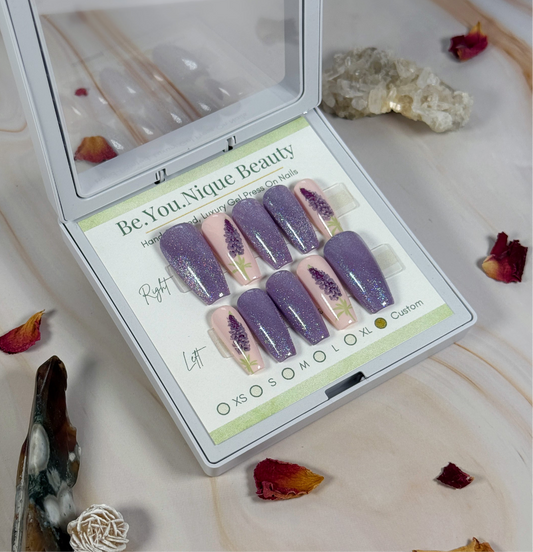 Set of purple and pink press-on nails in a 'Be.You.Nique Beauty' box on a decorative surface with crystals and flowers.