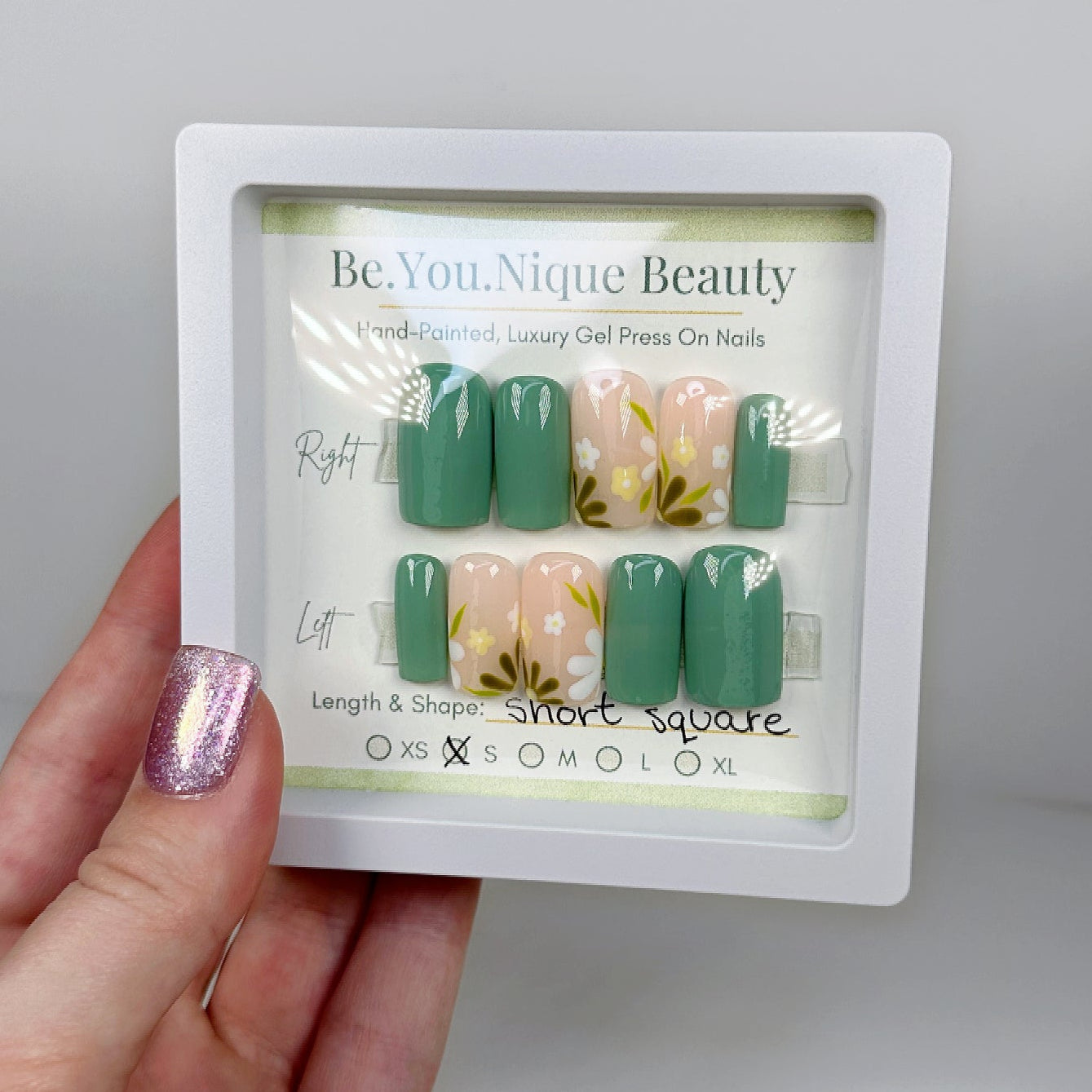 a set of 10 press on nails in size small. It is a short square set the thumb, pointer and ring fingers are a solid sage green colour while the middle and ring finger have a simple hand painted daisy floral design.