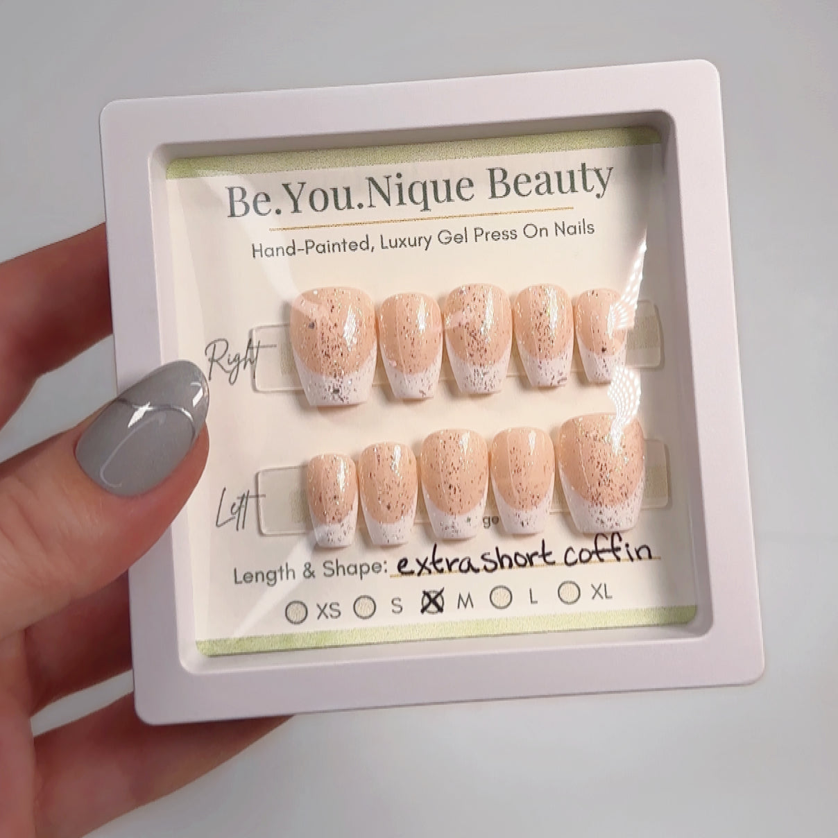 A set of press-on nails with a classic French tip design, featuring a soft nude base and a sparkling glitter top coat. The nails are neatly arranged in a box, ready for shipment, with a "Buy and Apply" label visible. The set includes multiple sizes for a custom fit and comes with adhesive tabs for easy application. The glitter top coat adds a touch of sparkle, making the nails perfect for a chic, elegant look.